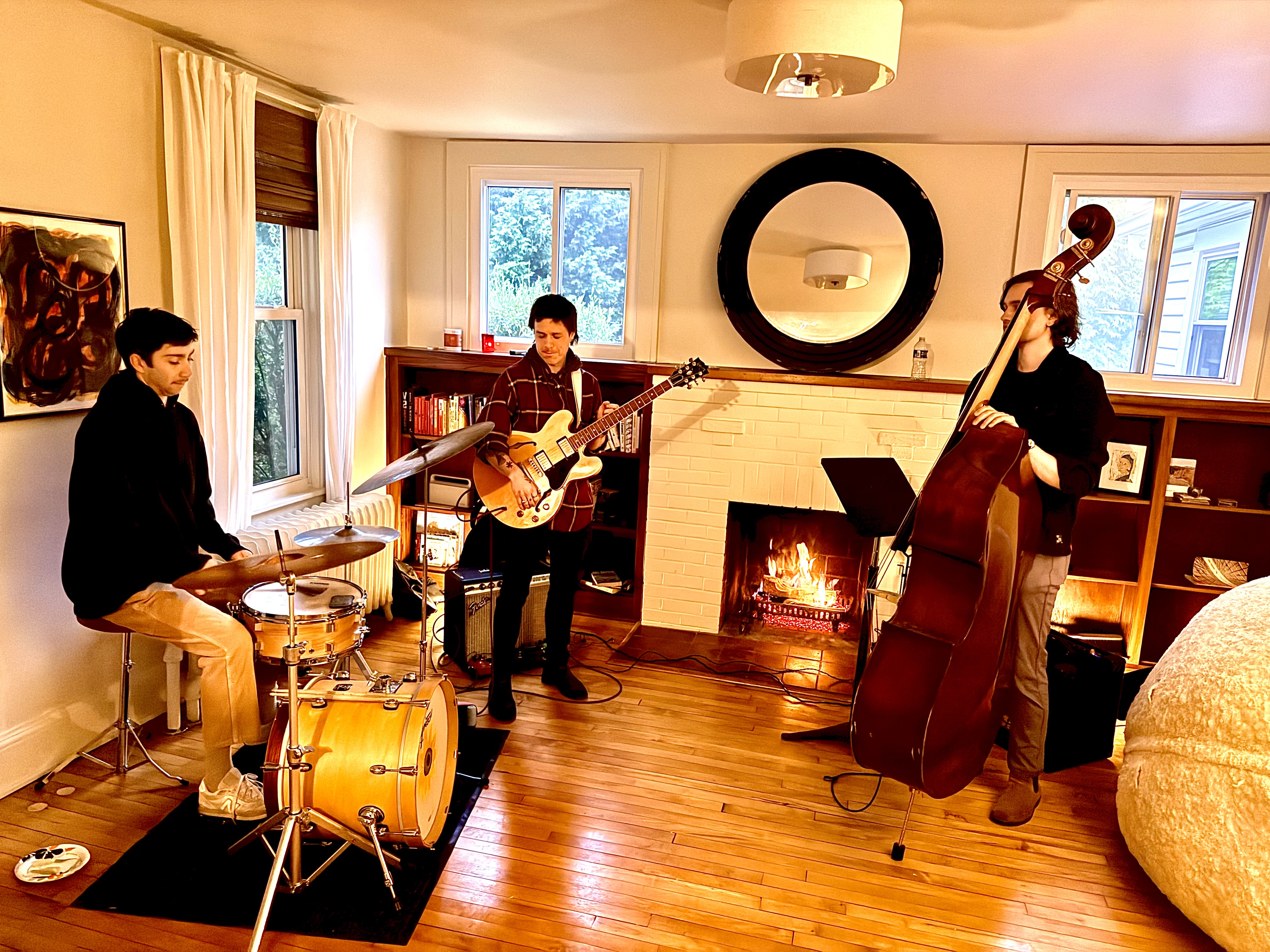 Jazz trio performing by the fireplace — drums, guitar, and upright bass in a cozy living room setting